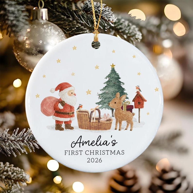 A round white ceramic Christmas ornament featuring a watercolor illustration of Santa Claus, a reindeer, a Christmas tree, and a birdhouse. The text reads Amelia's FIRST CHRISTMAS 2026.