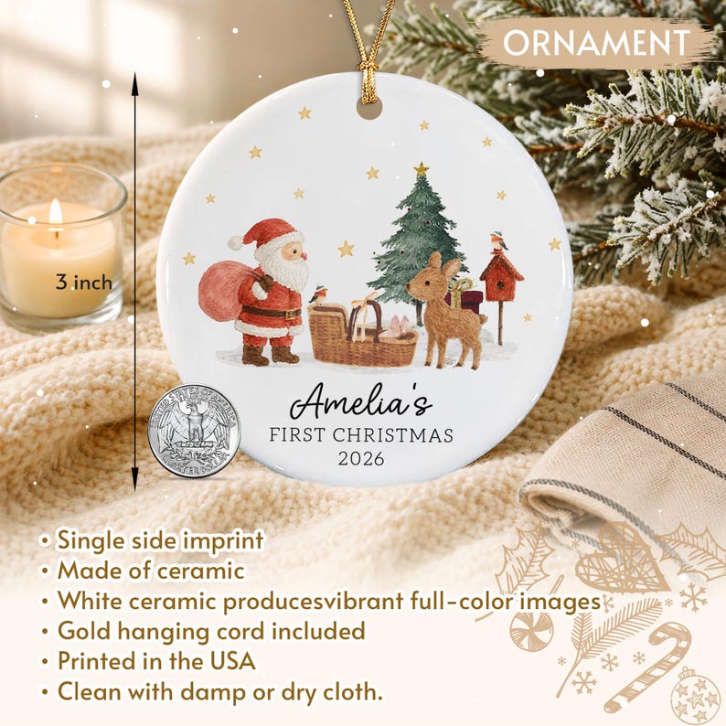 A round white ceramic ornament measuring 3 inches in diameter, featuring a gold hanging cord. The design includes a Santa Claus figure, a Christmas tree, a deer, and a birdhouse. Text reads: Amelia's FIRST CHRISTMAS 2026.