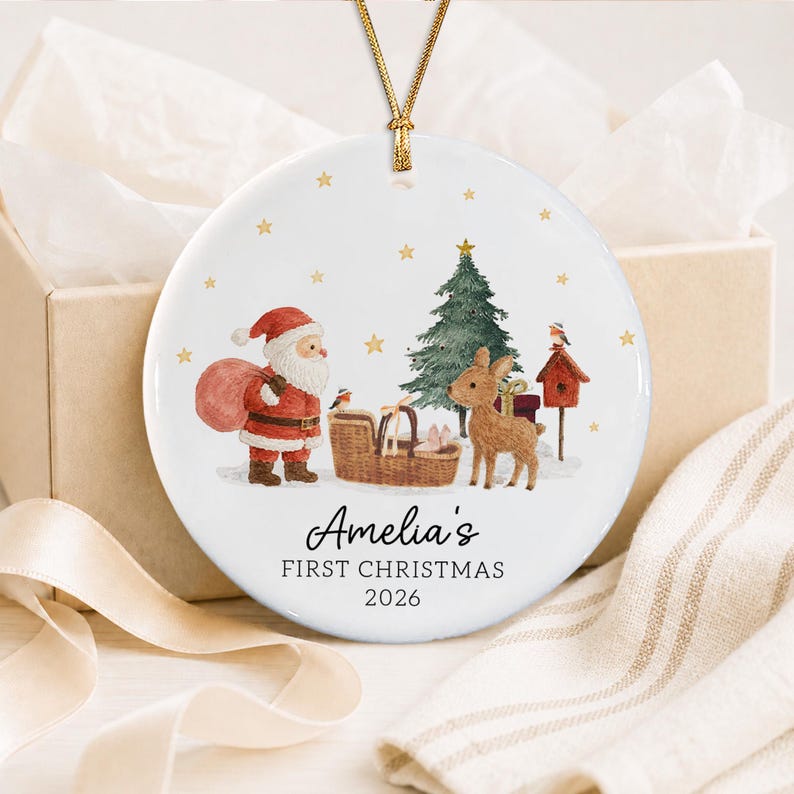 A round white ceramic Christmas ornament hanging from a gold cord. The design features a watercolor illustration of Santa Claus, a reindeer, a Christmas tree, and a birdhouse. Text reads: Amelia's FIRST CHRISTMAS 2026.