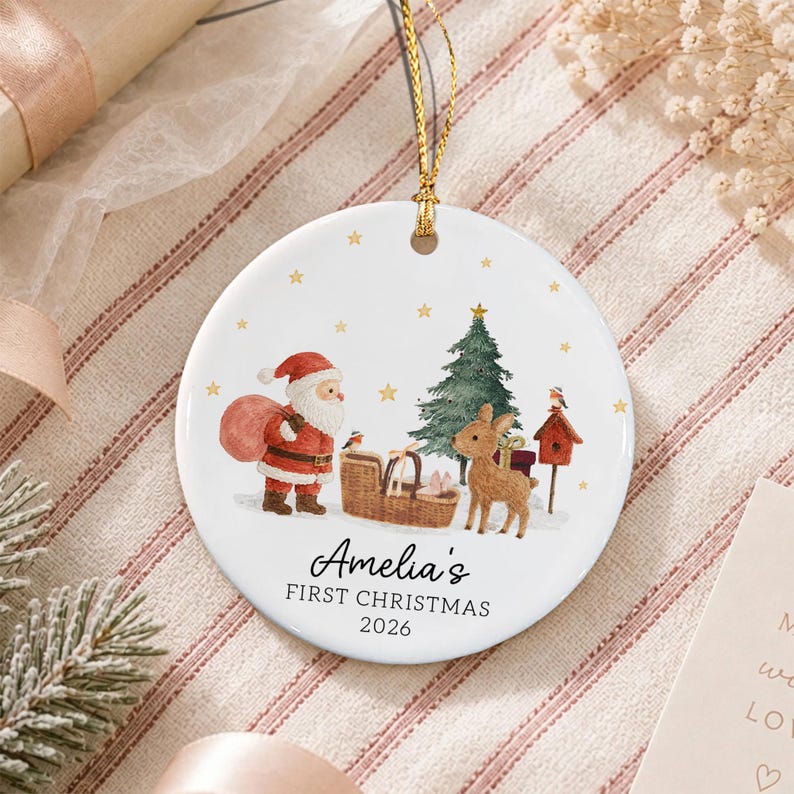 A round white ceramic ornament featuring a festive illustration of Santa Claus, a reindeer, a Christmas tree, and a birdhouse. The text reads Amelia's FIRST CHRISTMAS 2026.
