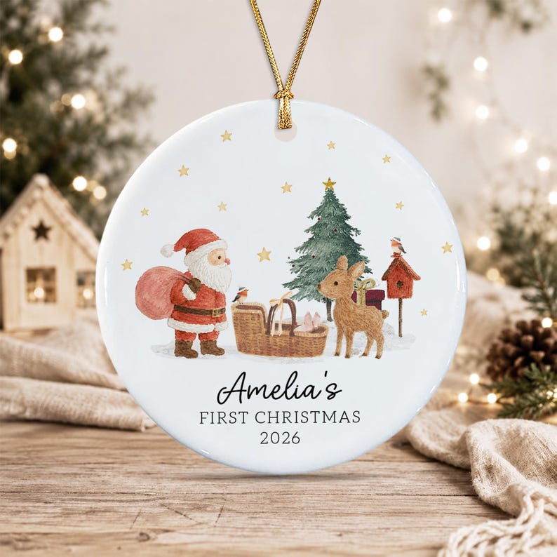A round white ceramic Christmas ornament featuring a watercolor illustration of Santa Claus, a reindeer, a Christmas tree, and a birdhouse. The text reads Amelia's FIRST CHRISTMAS 2026.