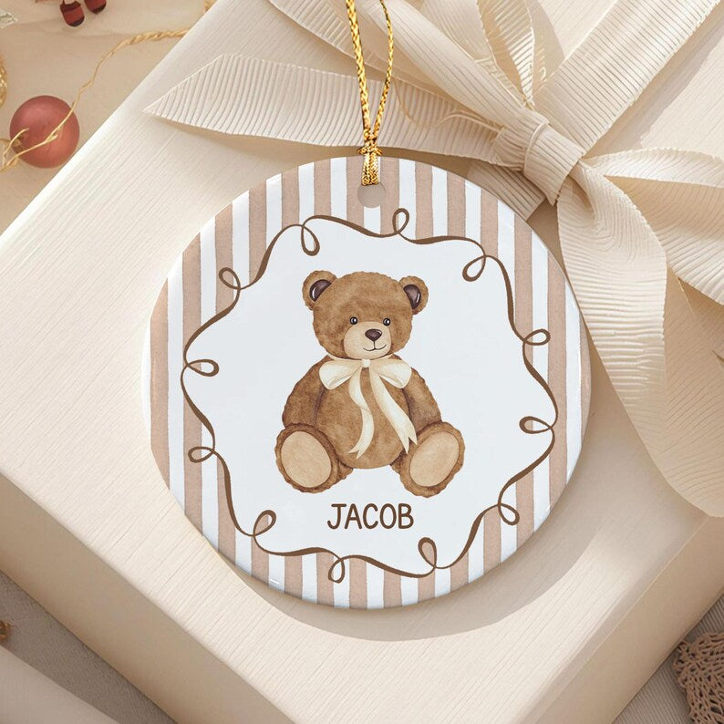 A round ceramic ornament featuring a brown teddy bear illustration with a cream-colored ribbon around its neck. The center is framed by a brown swirl border on a white background, surrounded by vertical beige and white stripes.