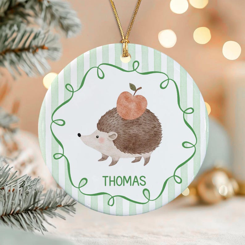 A round ceramic ornament featuring a light green and white vertical striped background. The center displays a brown hedgehog with a peach-colored apple on its back, enclosed by a green swirling border and the name THOMAS in green.