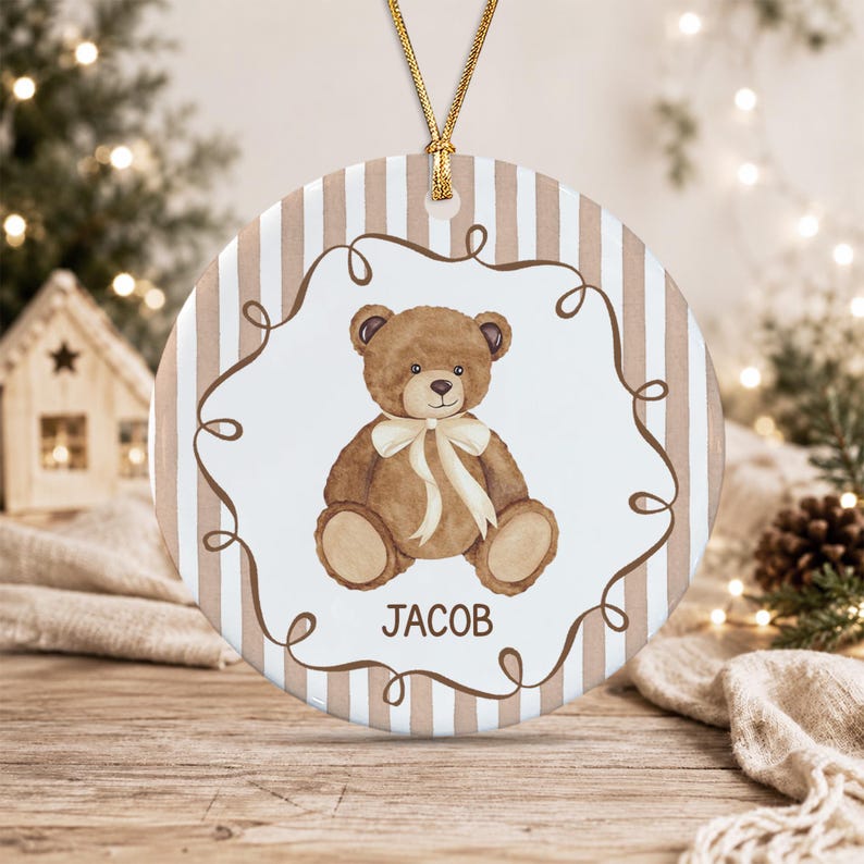 A round ceramic ornament featuring a brown teddy bear illustration with a cream-colored ribbon around its neck.