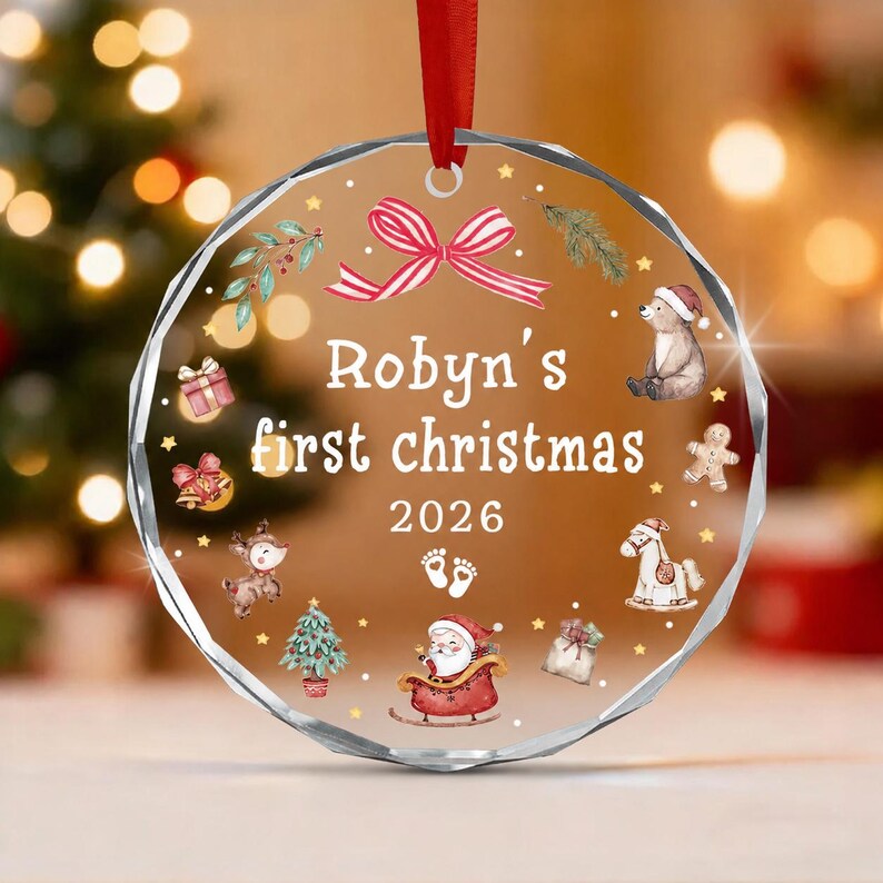 A round, clear acrylic ornament with faceted edges, hanging from a red ribbon. It features the text "Robyn's first christmas 2026" with illustrations of a red bow, a bear, a gingerbread man, a rocking horse, a gift, and Santa.