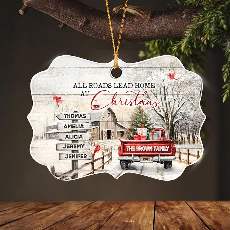 A decorative ornament with a scalloped edge featuring a winter scene with a red truck, a barn, and a signpost. The text reads ALL ROADS LEAD HOME AT CHRISTMAS, THE BROWN FAMILY, and 2025.