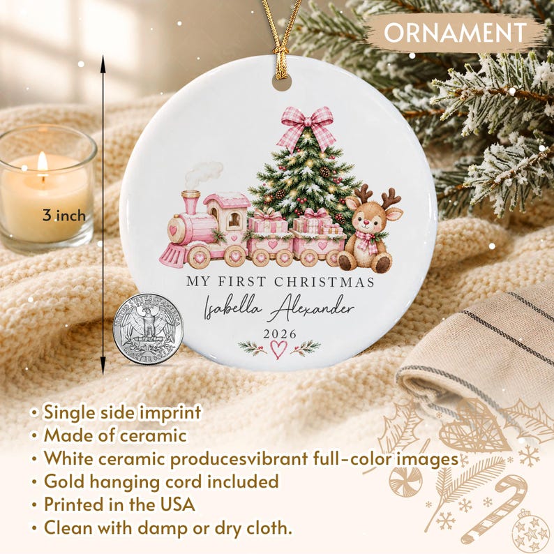 A round white ceramic ornament with a gold hanging cord, measuring 3 inches in diameter. The design features a pink train, a Christmas tree, and a reindeer, with the text "MY FIRST CHRISTMAS", "Isabella Alexander", and "2026".