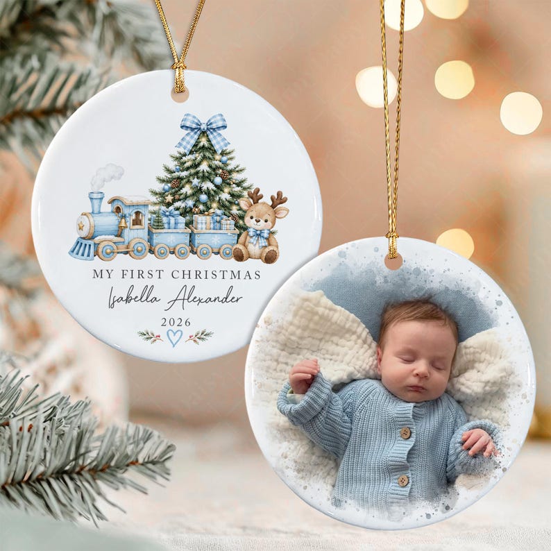 Two round white ceramic ornaments with gold hanging strings. One features a blue train, Christmas tree, reindeer, and the text "MY FIRST CHRISTMAS Isabella Alexander 2026".