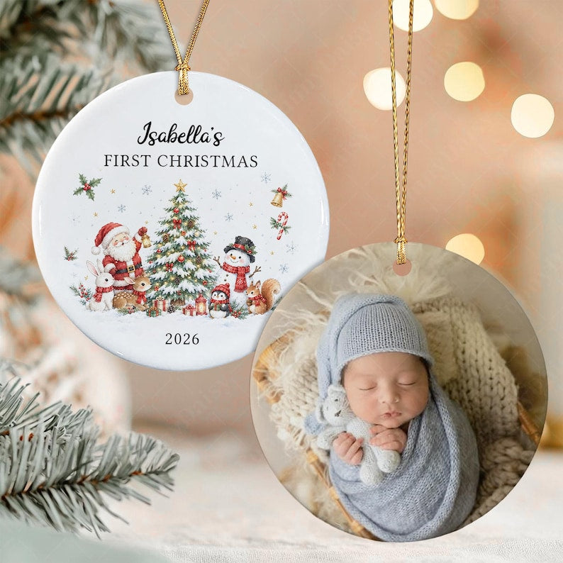 Two round ceramic ornaments with gold hanging strings. One ornament features a Christmas tree, Santa, a snowman, and the text Isabella's FIRST CHRISTMAS 2026.