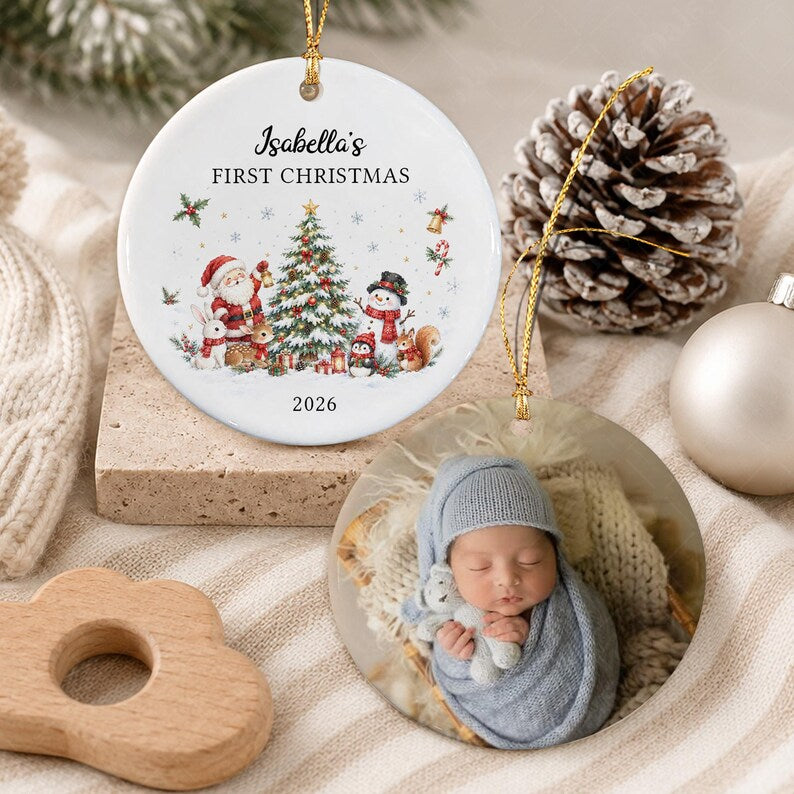 Two round ceramic ornaments with gold hanging strings. One features a Christmas tree, Santa, a snowman, and animals with the text Isabella's FIRST CHRISTMAS 2026.