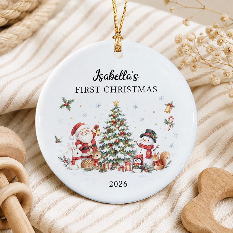 A round white ceramic Christmas ornament featuring a gold hanging string. The design includes the text Isabella's FIRST CHRISTMAS and 2026, with a central illustration of a decorated tree, Santa Claus, a snowman, and animals.