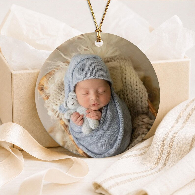 A circular ceramic ornament featuring a photograph of a newborn baby wrapped in a light blue knitted blanket and matching cap, holding a small gray plush toy. The ornament hangs from a gold cord and is displayed in a gift box.