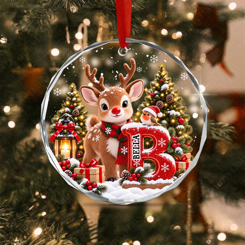 A round, faceted glass Christmas ornament featuring a cartoon reindeer wearing a red and green scarf, a small bird, a lit lantern, and a large red letter B with the name BELLA printed on it, all set against a snowy forest.