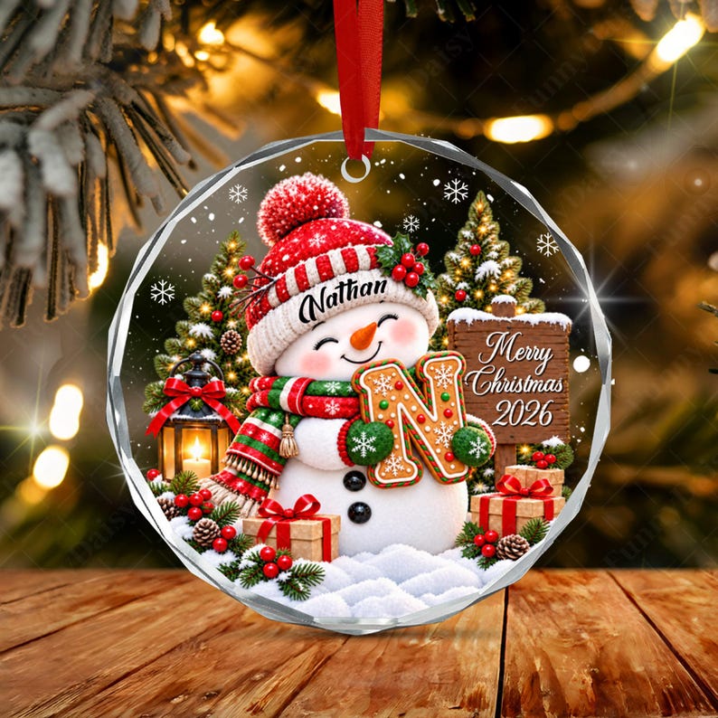 A round, clear acrylic Christmas ornament featuring a snowman wearing a red hat with the name Nathan, a red and green scarf, and holding a large letter N. The design includes a wooden sign reading Merry Christmas 2026.