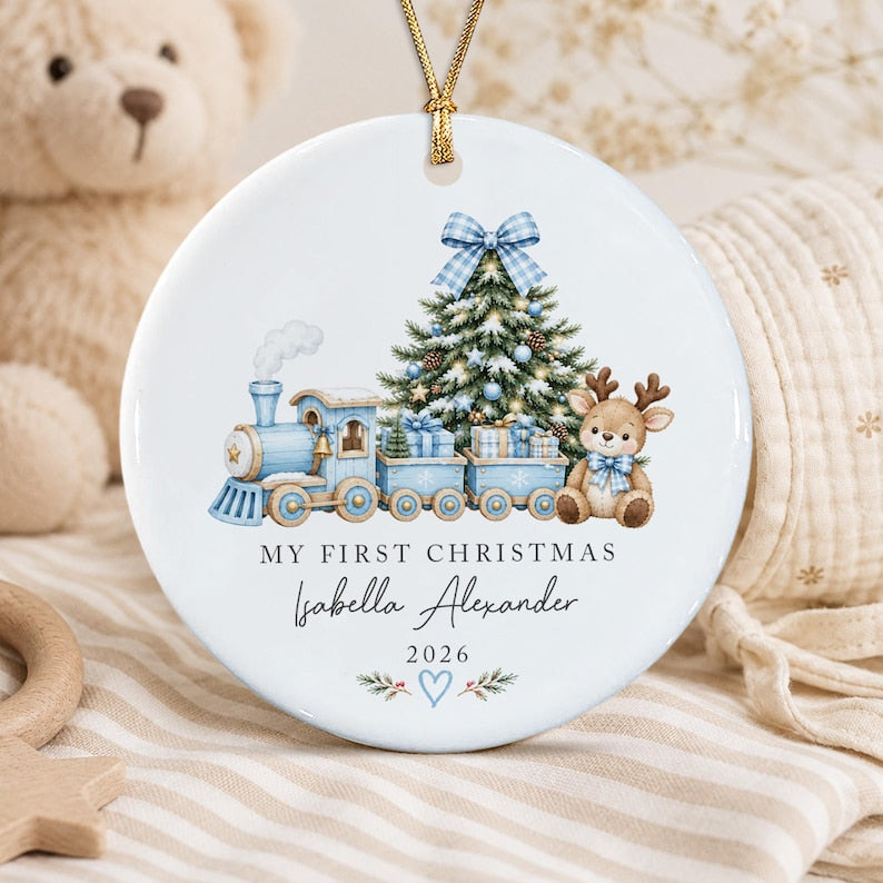 A round white ceramic ornament featuring a blue train, a decorated Christmas tree, and a reindeer. The text reads MY FIRST CHRISTMAS, Isabella Alexander, and 2026, with a blue heart and pine branches at the bottom.