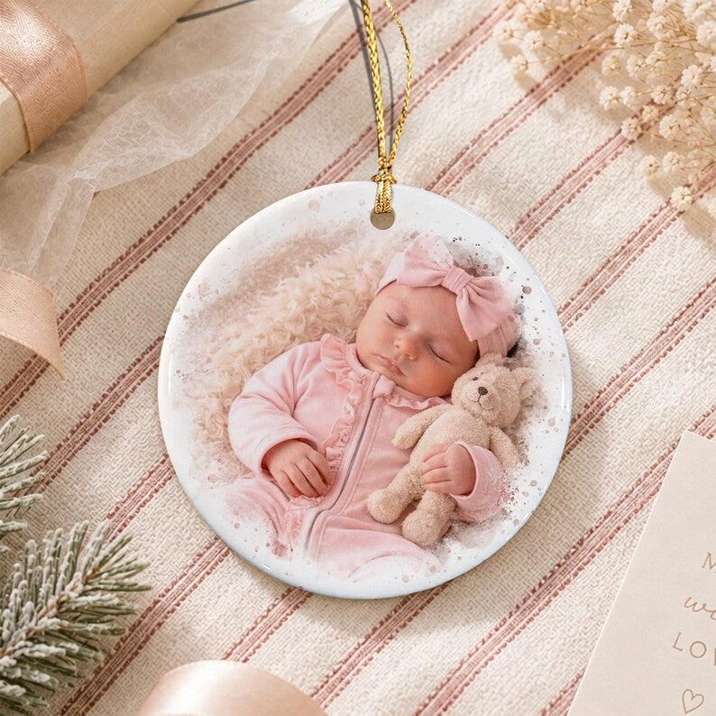 A round white ceramic ornament featuring a printed photograph of a sleeping infant wearing a light pink outfit and a matching headband with a bow. The infant is holding a small tan teddy bear. A gold string is attached to the top.