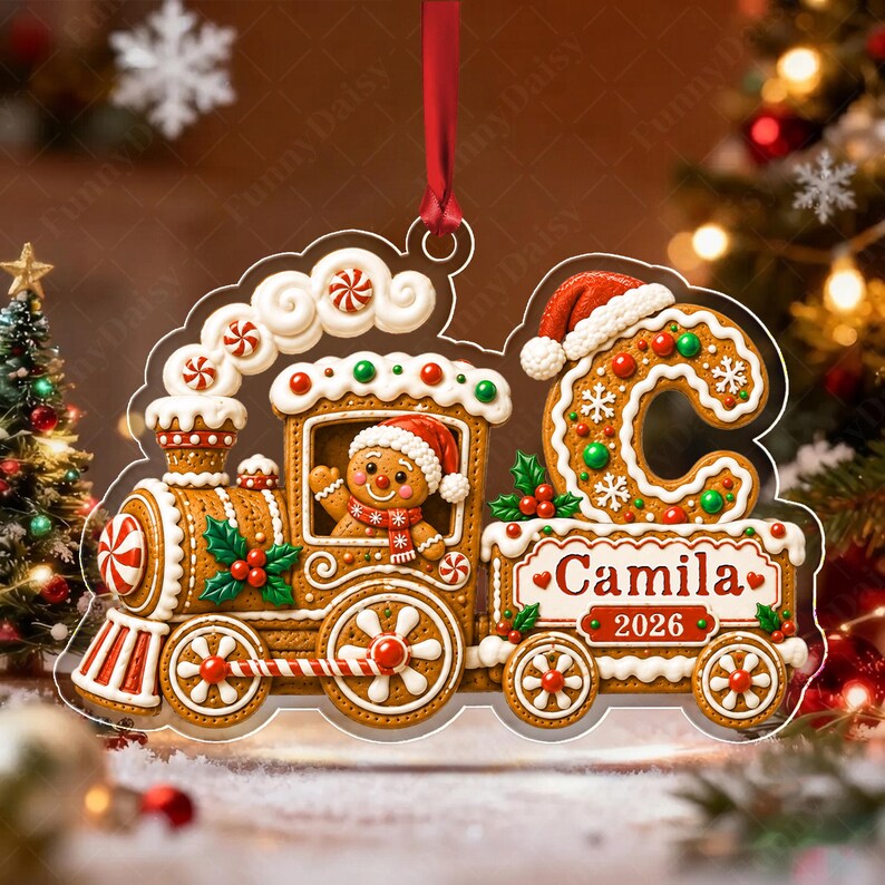 A gingerbread train Christmas ornament featuring a gingerbread person in a Santa hat inside the engine, a large letter C, and a carriage labeled with the name Camila and the year 2026, all suspended by a red ribbon.