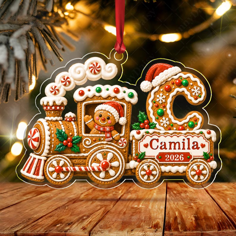 A gingerbread train Christmas ornament featuring a gingerbread person in a Santa hat, a large letter C, and the text Camila 2026. The ornament is decorated with white icing, red and green accents, and hangs from a red ribbon.