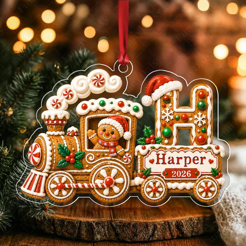 A gingerbread train Christmas ornament featuring a gingerbread person in the engine, a large letter H with a Santa hat, and a carriage labeled Harper 2026. The design includes white icing, red and green candies, and a red ribbon.