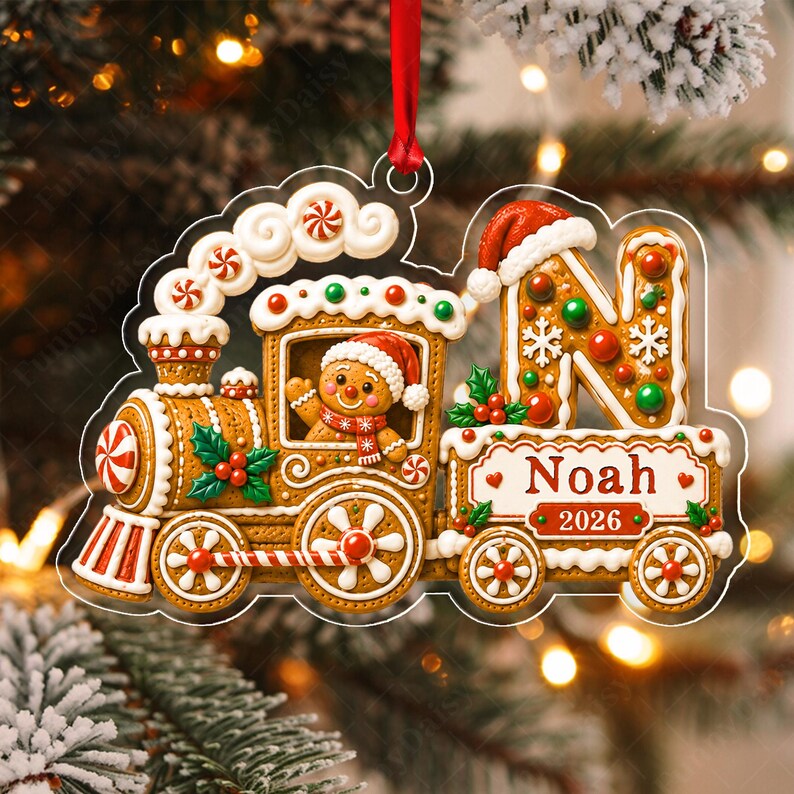 A gingerbread train Christmas ornament hanging from a red ribbon. The train features a gingerbread person in the cabin, a large letter N with a Santa hat, and a banner displaying the name Noah and the year 2026 in red text.