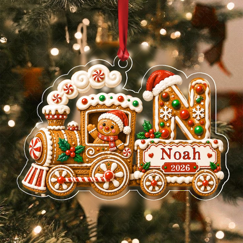 A gingerbread train Christmas ornament featuring a gingerbread person in a Santa hat, a large letter N with a Santa hat, and a white banner with the text Noah and 2026. The ornament is decorated with red and green accents.