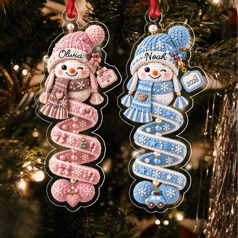 Two flat acrylic Christmas ornaments featuring crocheted-style snowman designs. One is pink with the name Olivia and the year 2026, the other is blue with the name Noah and the year 2026. Both have spiral tails with small charms.