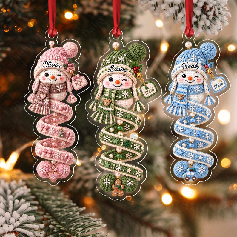 Three flat acrylic Christmas ornaments featuring crocheted snowman designs in pink, green, and blue. Each snowman wears a matching hat and scarf, with names Olivia, Liam, and Noah printed above.
