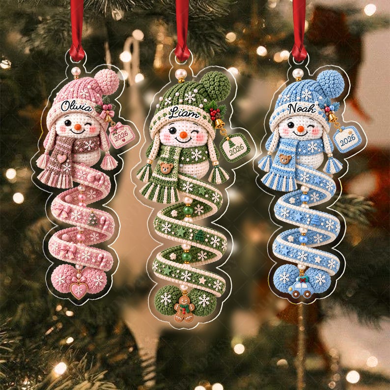 Three hanging ornaments featuring crocheted snowmen in pink, green, and blue. Each snowman wears a matching hat and scarf, with a spiral tail decorated with small charms.