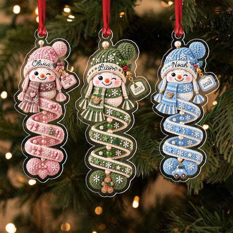 Three acrylic Christmas ornaments featuring crocheted-style snowmen in pink, green, and blue. Each snowman wears a matching hat and scarf, with names Olivia, Liam, and Noah printed above. A spiral tail hangs below with 2026 tags.