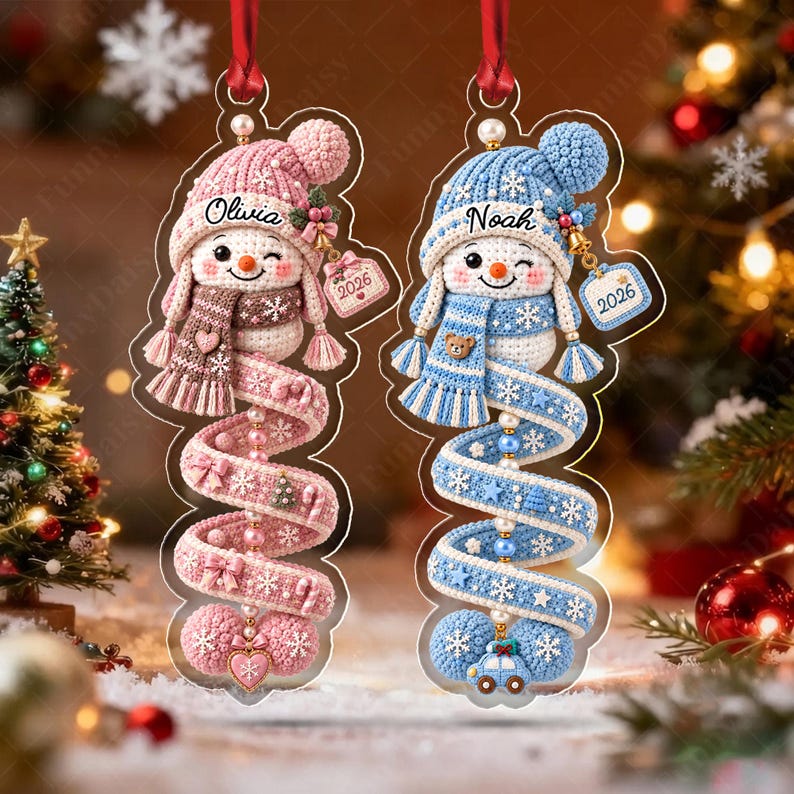 Two hanging acrylic ornaments featuring crocheted snowman designs. One snowman is pink with the name Olivia and the year 2026, while the other is blue with the name Noah and the year 2026. Both have spiral tails with beads.