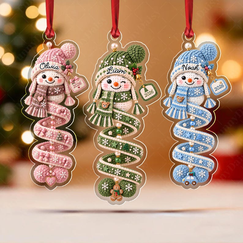 Three acrylic Christmas ornaments featuring crocheted snowman designs in pink, green, and blue. Each snowman wears a matching hat and scarf, with personalized names Olivia, Liam, and Noah, and a small tag displaying the year 2026.