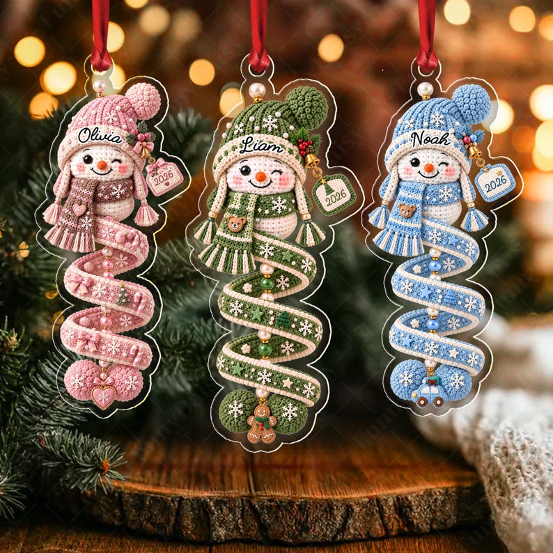 Three hanging Christmas ornaments featuring crocheted-style snowmen in pink, green, and blue. Each snowman wears a matching hat and scarf, with names Olivia, Liam, and Noah printed on the hats. Each ornament includes a 2026 tag.