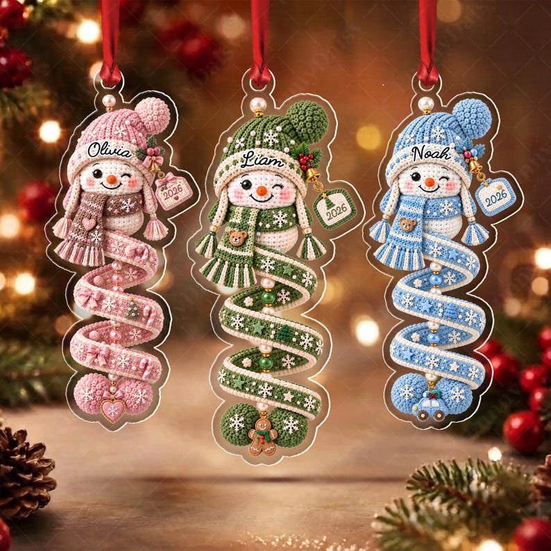 Three hanging ornaments featuring crocheted snowmen in pink, green, and blue. Each snowman wears a matching hat and scarf, with a spiral tail decorated with small charms.