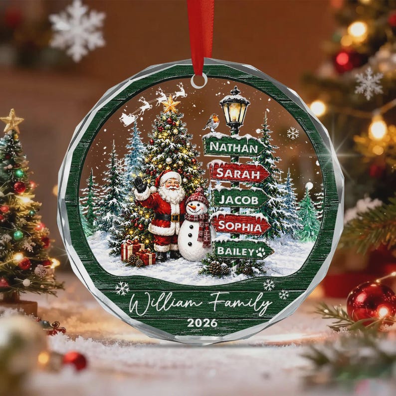 A round, clear acrylic Christmas ornament featuring a printed winter scene with Santa Claus, a snowman, and a street sign listing the names Nathan, Sarah, Jacob, Sophia, and Bailey.