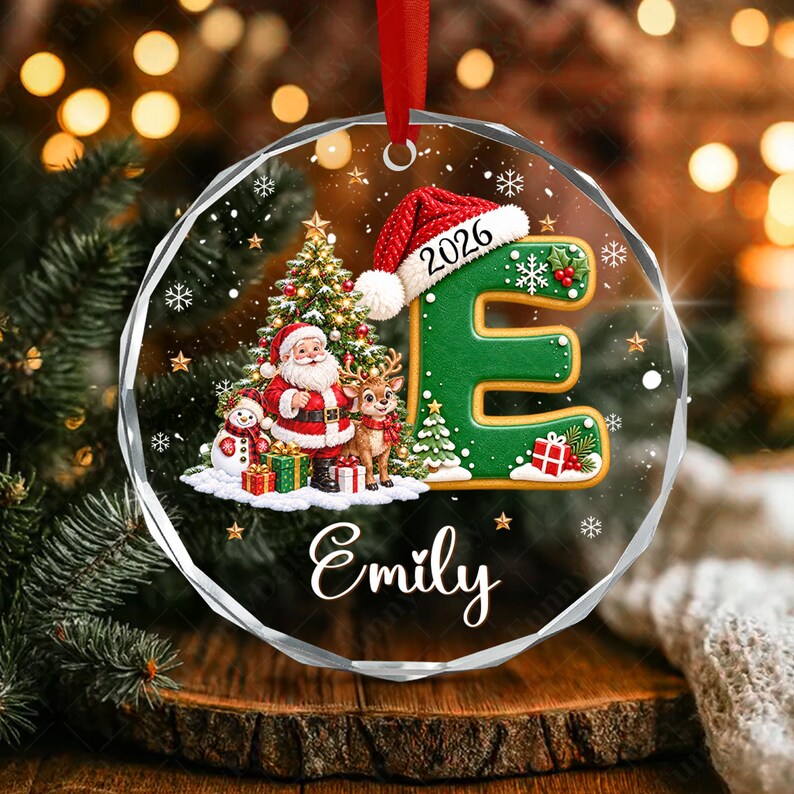 A round, clear acrylic Christmas ornament featuring a green letter E wearing a red Santa hat with the year 2026. The design includes a Santa Claus, a reindeer, a snowman, and gifts under a tree, with the name Emily printed below.