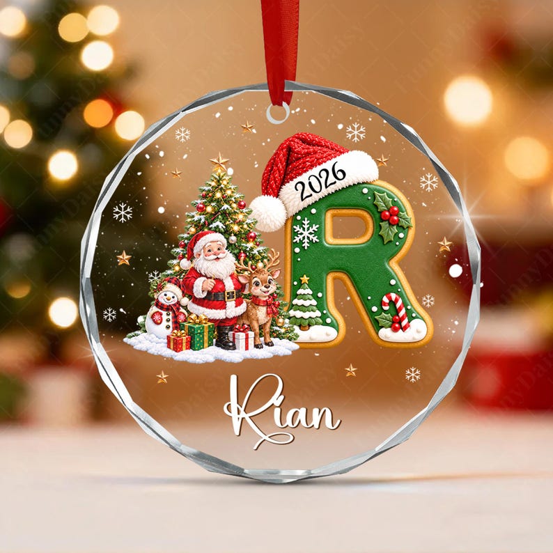 A round, faceted glass Christmas ornament featuring a green letter R wearing a red Santa hat with the year 2026. The design includes Santa Claus, a snowman, a reindeer, a Christmas tree, and the name Rian in white script text.