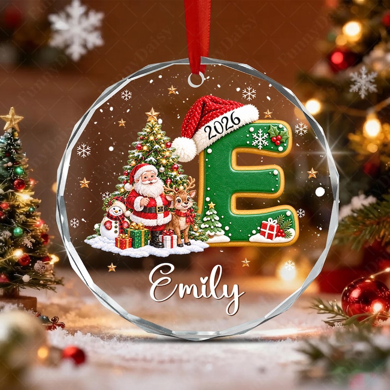 A circular clear acrylic Christmas ornament featuring a green letter E wearing a Santa hat with the year 2026, a Santa Claus figure, a reindeer, a snowman, and gifts.