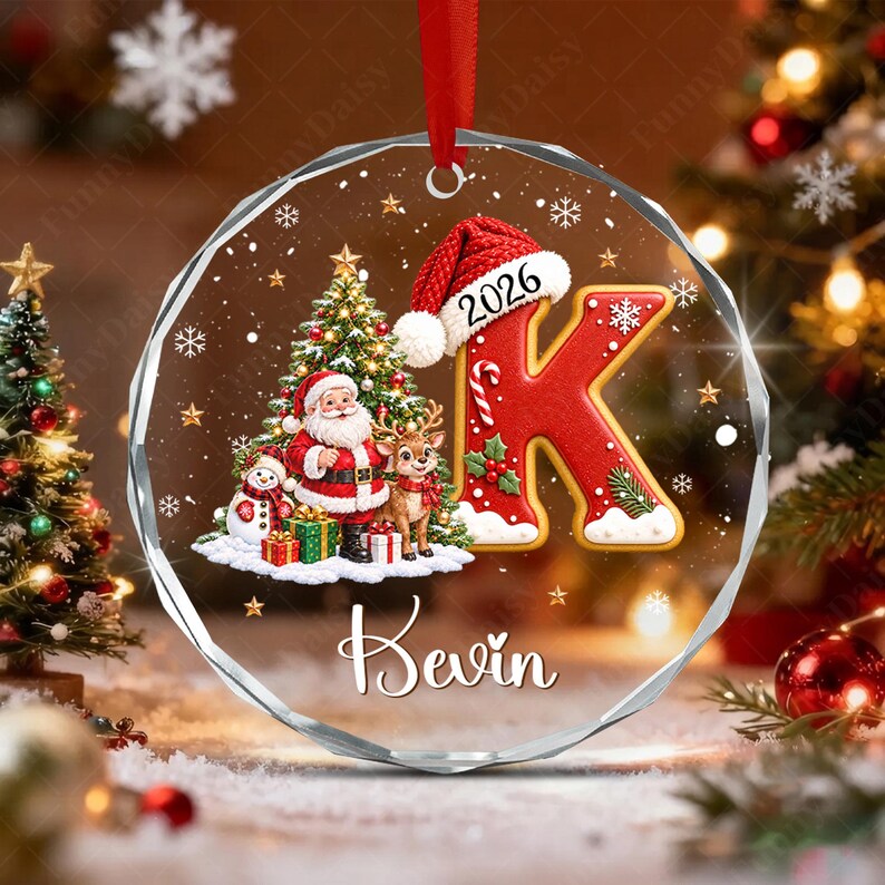 A round, clear acrylic Christmas ornament featuring a red letter K wearing a Santa hat with the year 2026, a Santa Claus figure, a reindeer, a snowman, and gifts.