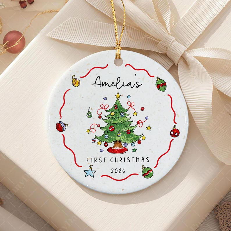 A round white ceramic ornament featuring a hand-drawn green Christmas tree with colorful baubles and stars. The text reads Amelia's FIRST CHRISTMAS 2026.