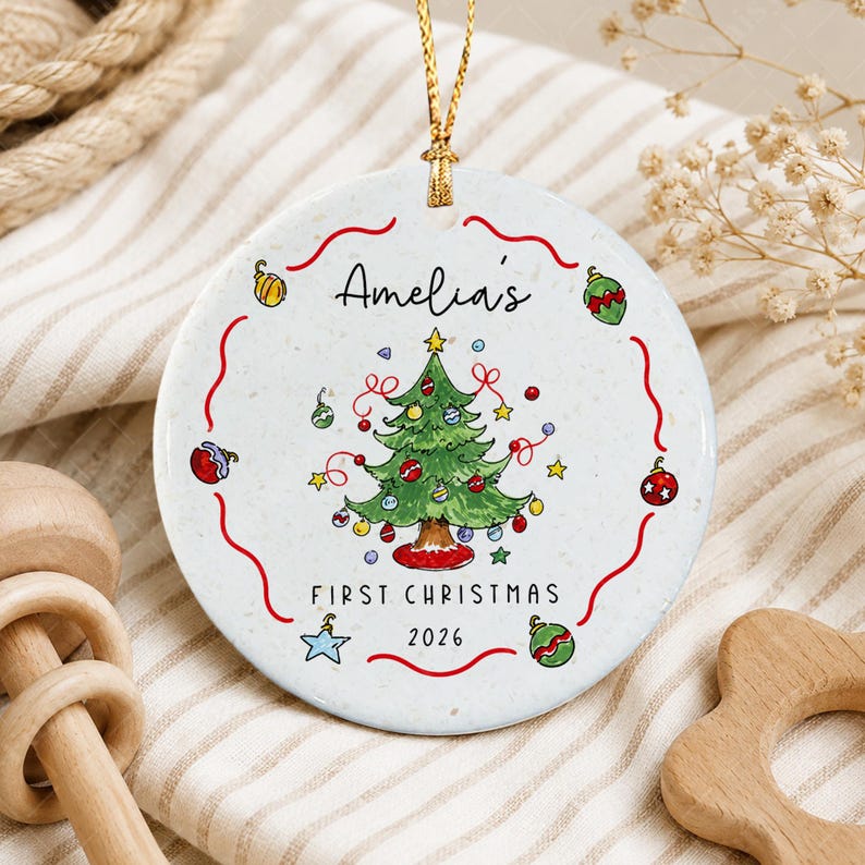 A round white ceramic ornament featuring a hand-drawn green Christmas tree with colorful baubles and stars. The text reads Amelia's FIRST CHRISTMAS 2026.