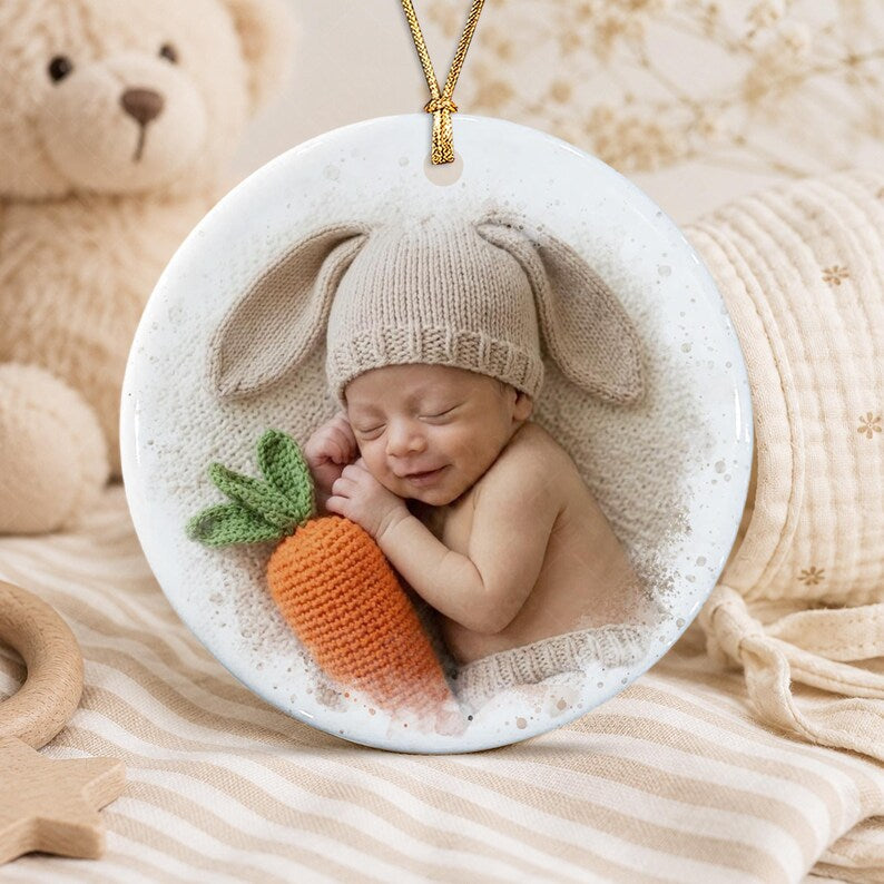 A round white ceramic ornament featuring a printed image of a sleeping infant wearing a beige knit hat with long bunny ears. The infant is resting next to a small, textured orange crocheted carrot with green leaves.