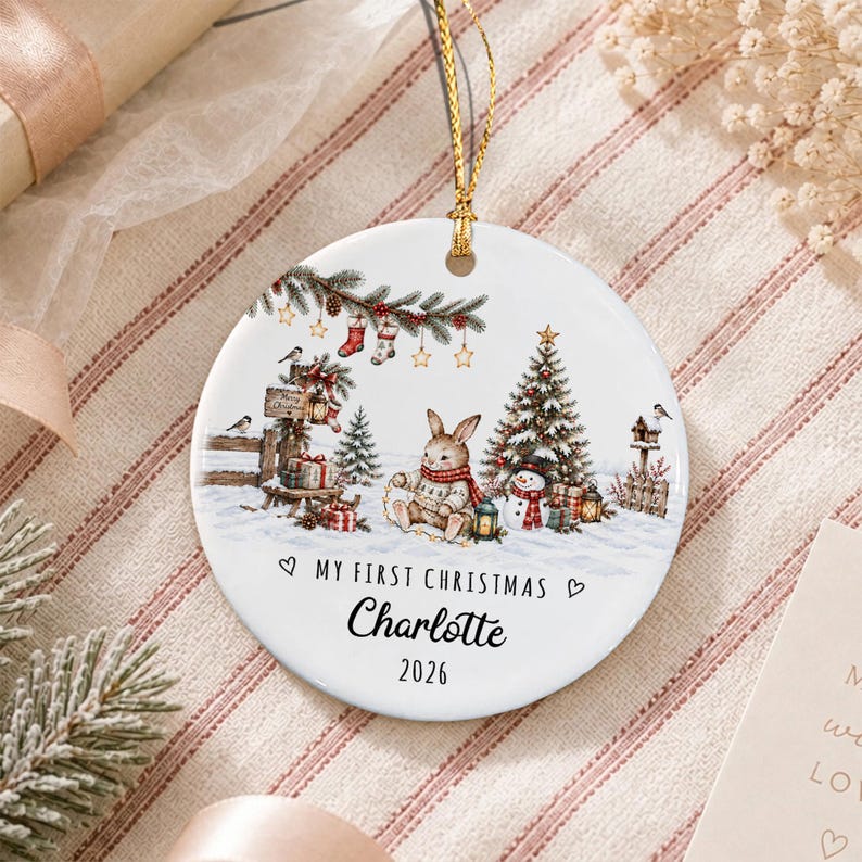 A round white ceramic ornament featuring a festive winter scene with a rabbit, a Christmas tree, a snowman, and hanging stockings. The text reads My First Christmas, Charlotte, and 2026. A gold string is attached for hanging.