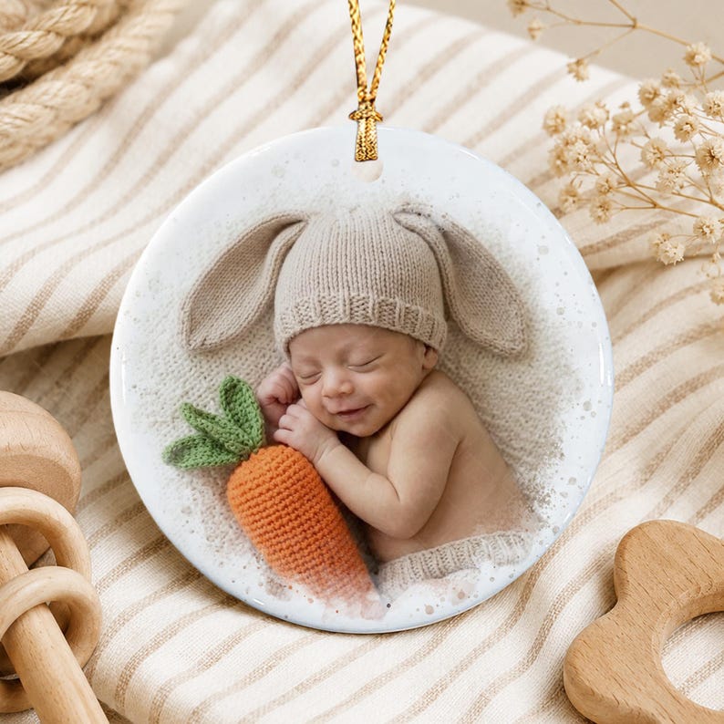 A round ceramic ornament featuring a printed image of a sleeping infant wearing a beige knit hat with bunny ears. The infant is holding a small, textured orange crocheted carrot with green leaves.
