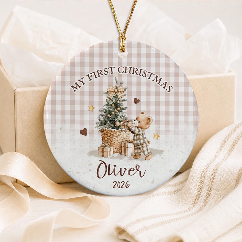 A round ceramic Christmas ornament featuring a light brown gingham background. The design includes a decorated Christmas tree, a teddy bear in pajamas, and the text "MY FIRST CHRISTMAS", "Oliver", and "2026".