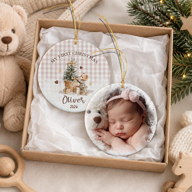Two round ceramic Christmas ornaments with gold hanging strings. One ornament features a beige gingham pattern, a teddy bear illustration, and the text "MY FIRST CHRISTMAS Oliver 2026".