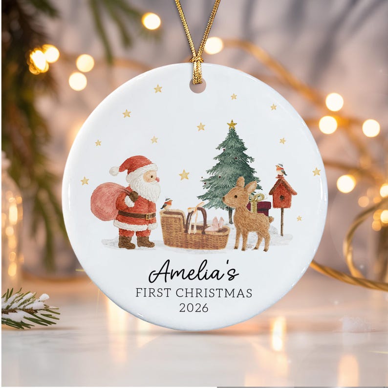 A round white ceramic ornament with a gold string featuring a watercolor illustration of Santa Claus, a reindeer, a Christmas tree, and a birdhouse.