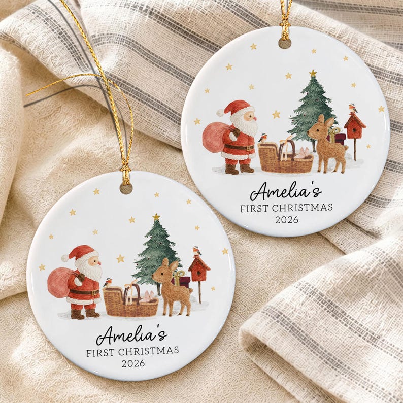 Two round white ceramic ornaments with gold hanging strings. Each features a watercolor illustration of Santa Claus, a Christmas tree, a basket, a birdhouse, and a deer. Text reads: Amelia's FIRST CHRISTMAS 2026.