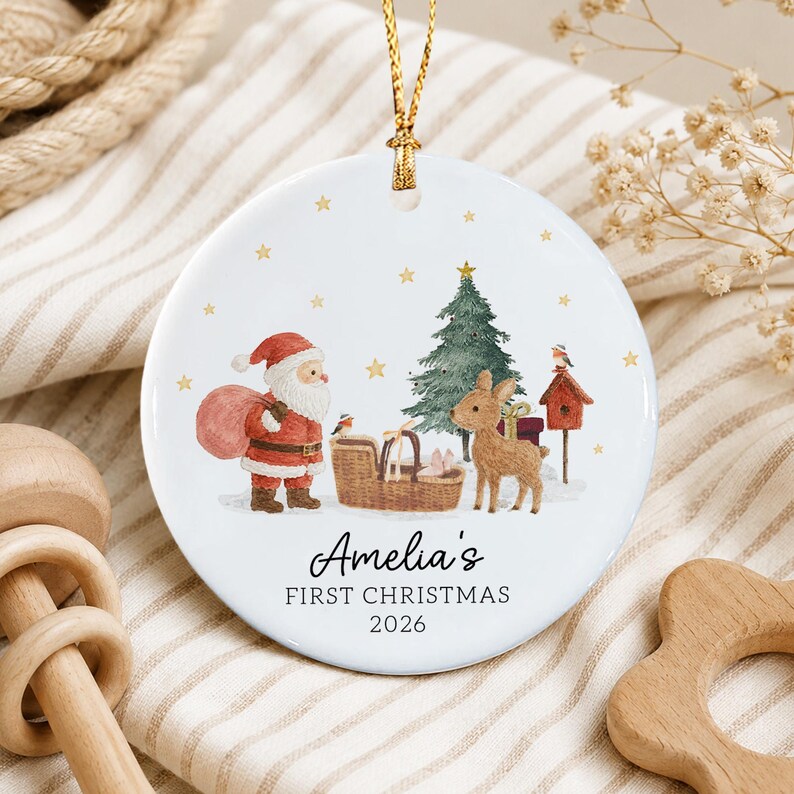 A round white ceramic Christmas ornament featuring a watercolor illustration of Santa Claus, a reindeer, a Christmas tree, and a birdhouse. The text reads Amelia's FIRST CHRISTMAS 2026.