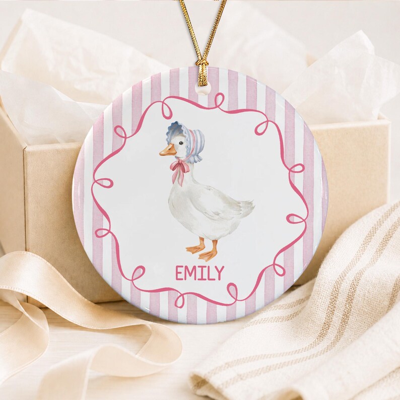 A round ceramic ornament featuring a white duck wearing a blue and white striped bonnet with a pink bow.