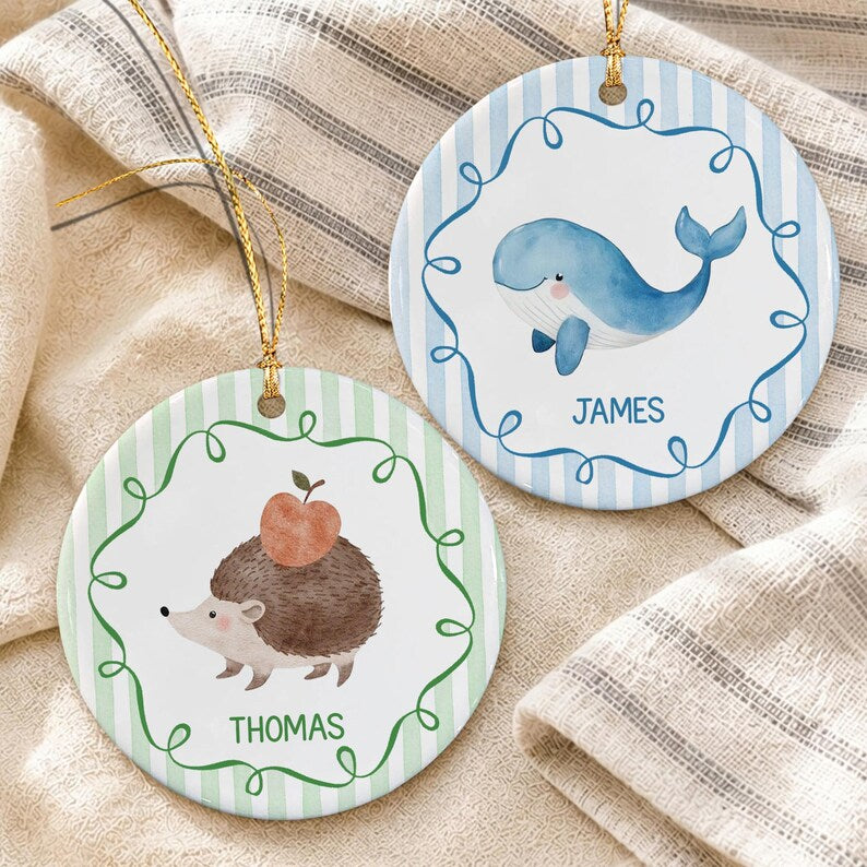 Two round ceramic ornaments with gold hanging strings. One features a blue whale and the name JAMES on a blue striped background. The other shows a brown hedgehog with an apple and the name THOMAS on a green striped background.